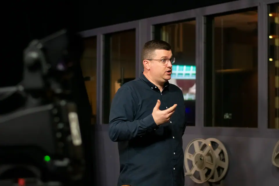 Production Stills — Presenter speaking on a broadcast studio set, a studio camera and film reels framing the shot