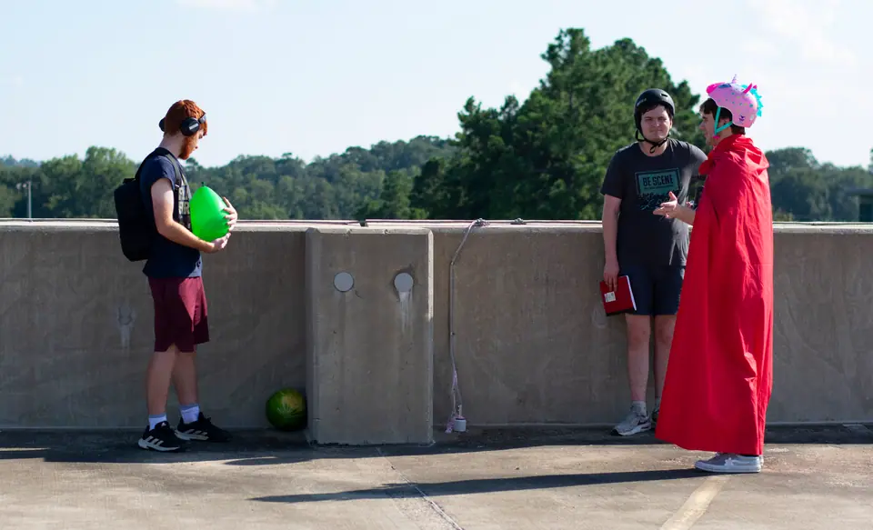 Two actors rehearsing on a rooftop, one in a red cape, crew watching from the side