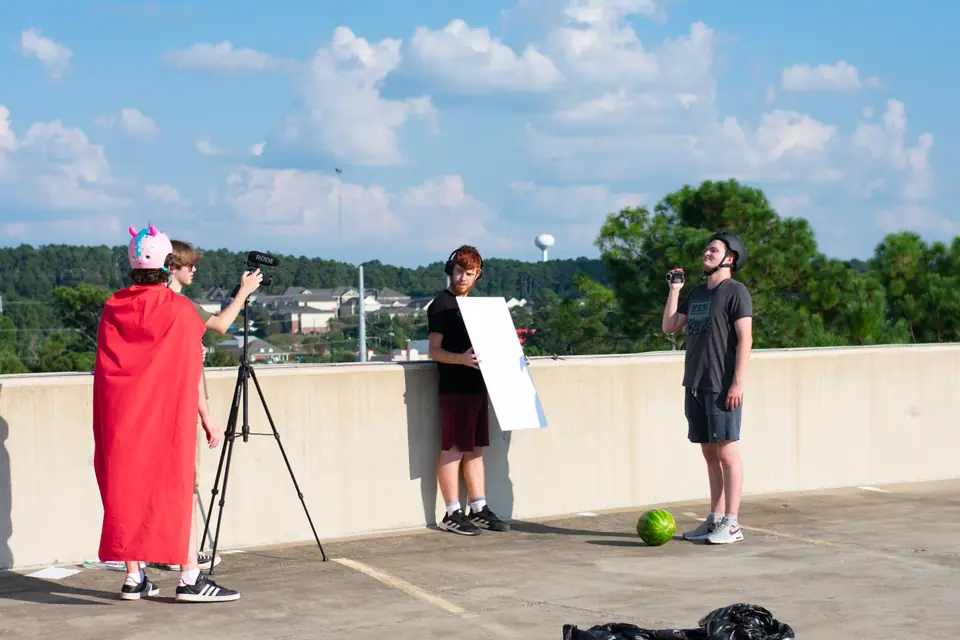 Student film crew on a parking garage roof, one holding a bounce board beside a tripod