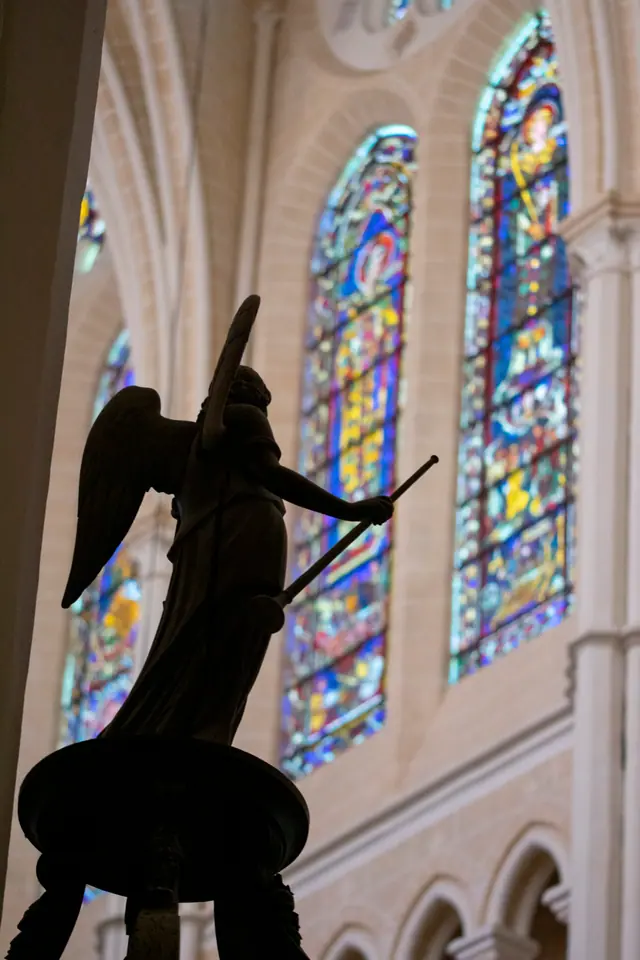 Angel sculpture holding a staff, framed by stained glass and pale vaulting behind