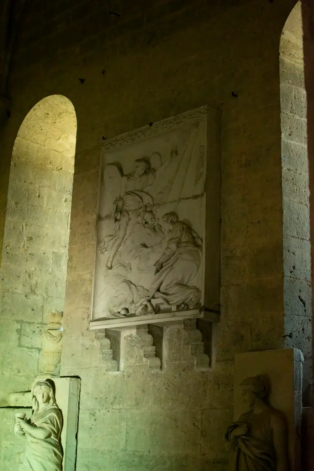 Carved relief panel set into a warm-lit stone wall, a standing figure beside it
