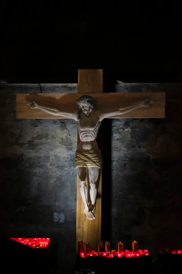 Crucifix mounted on a dark stone wall above a rack of lit red votive candles