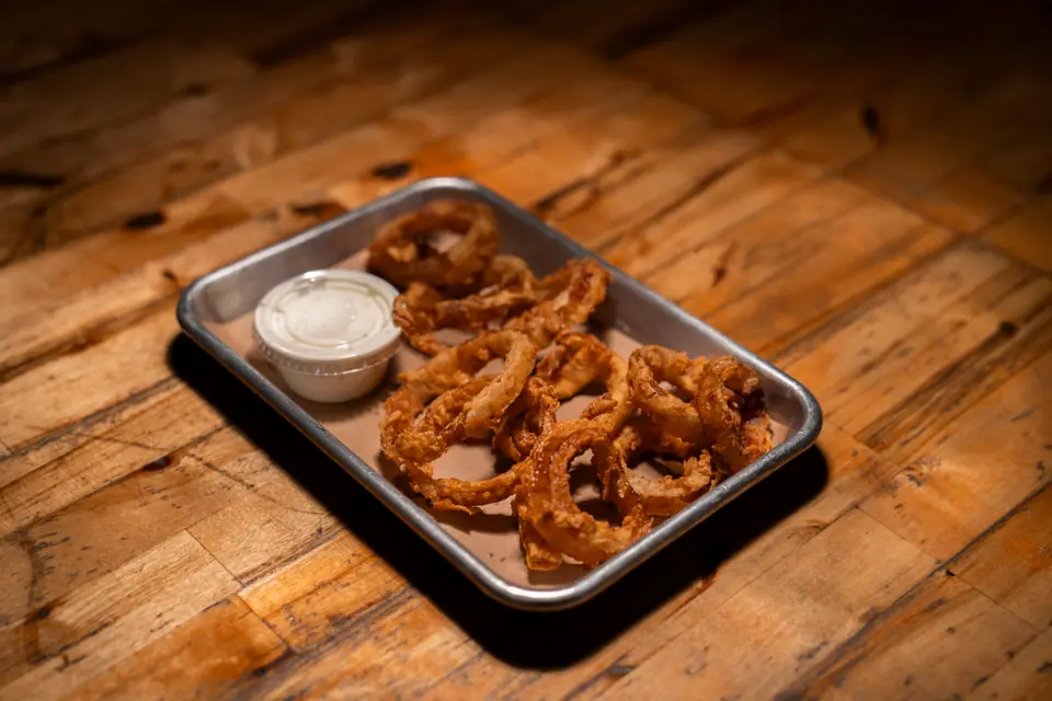 Basket of onion rings with a cup of dipping sauce on a metal tray, on a rustic wood table