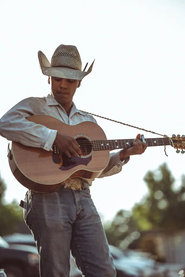 Young musician in a straw cowboy hat playing an acoustic guitar outdoors in backlight