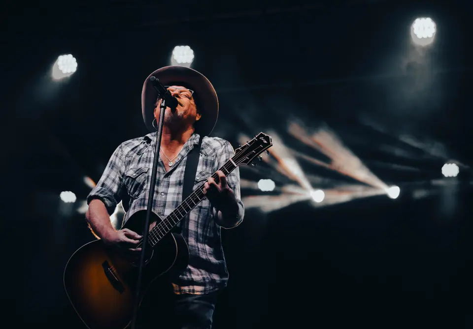 Country singer in a wide-brimmed hat at the microphone with an acoustic guitar, light beams behind