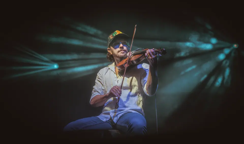 Fiddler kneeling and playing under blue stage light with lens flare across the frame