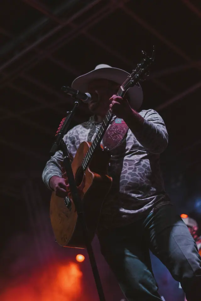 Guitarist in a patterned shirt and hat performing under deep red stage light
