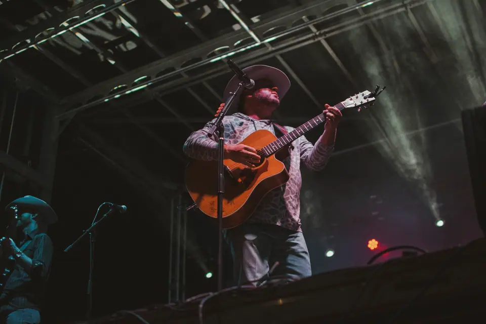 Guitarist in a wide-brimmed hat playing at the front of the stage through drifting smoke