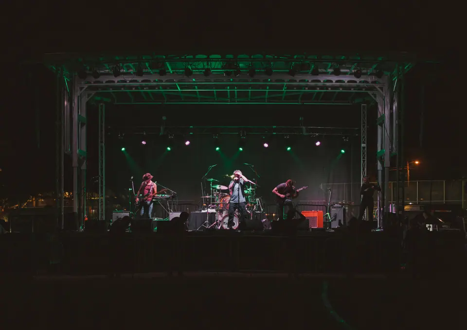 Wide view of a band on an outdoor stage at night under green light, silhouetted crowd in front