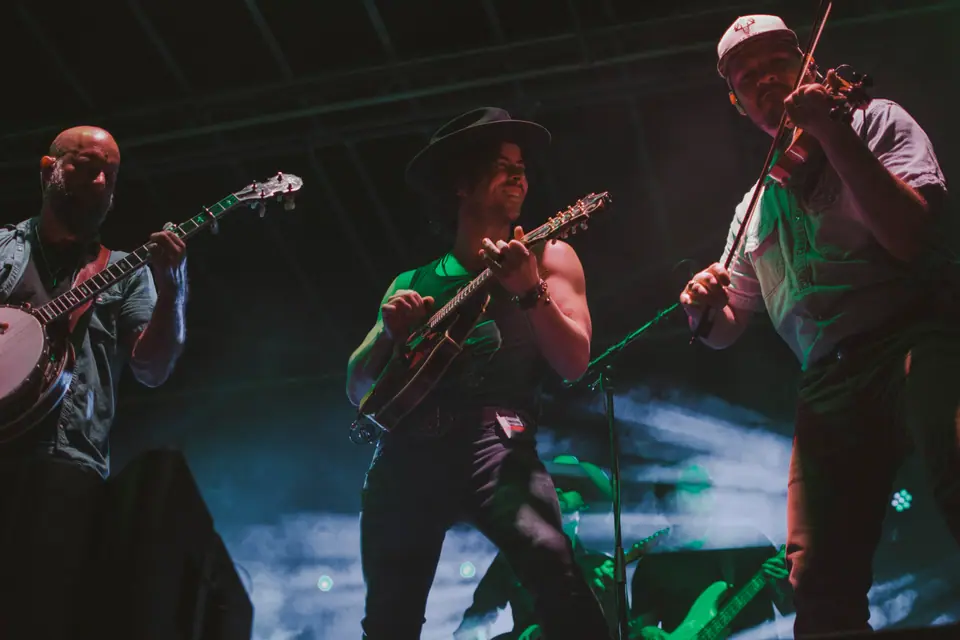 Three-piece string band on stage under green light, playing banjo, guitar, and fiddle