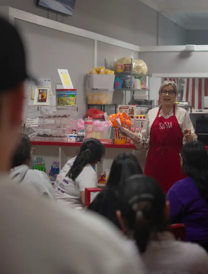 Presenter in a red apron speaking to a seated audience in a small shop