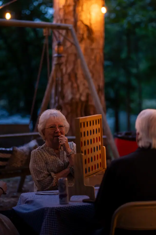 Older guest listening at a table at dusk, lit by a nearby fire