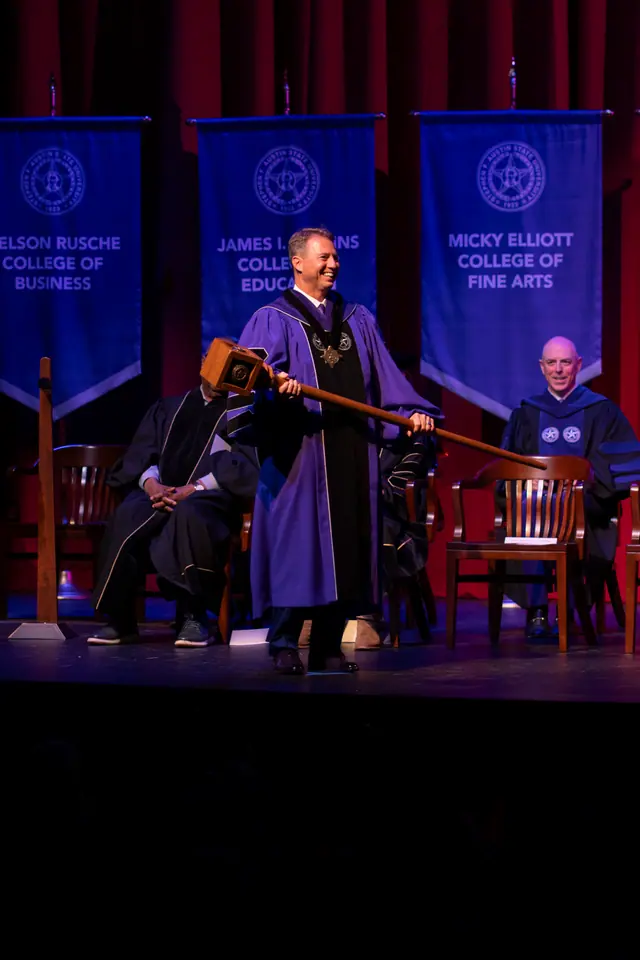 Official in academic regalia carrying a ceremonial mace across the stage in front of college banners