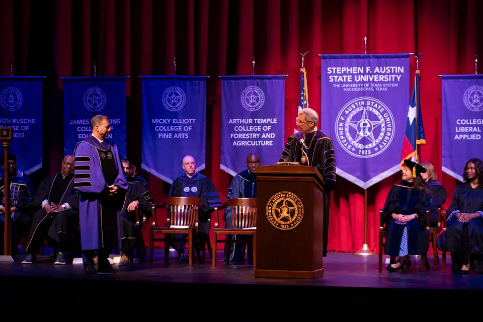 University investiture ceremony on stage, faculty in academic regalia beside college banners and a seal-fronted podium