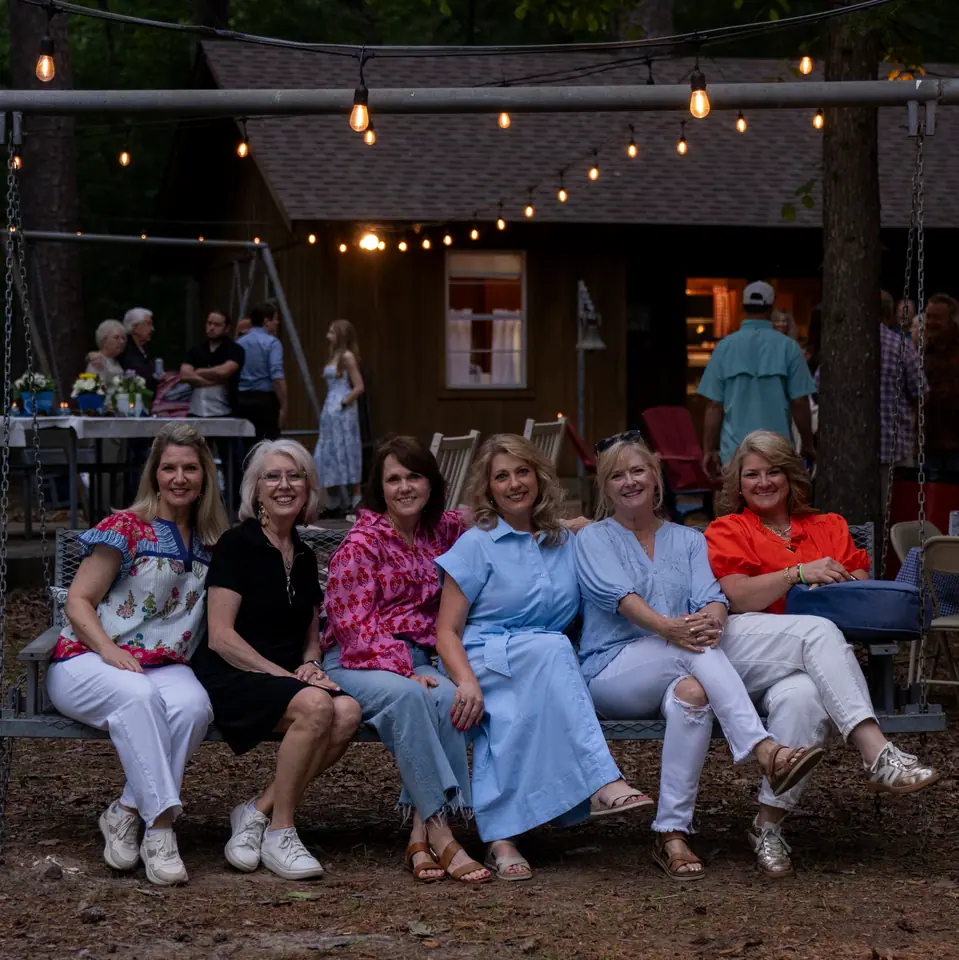 Six women seated together on a bench outdoors, string lights and pine trees behind them