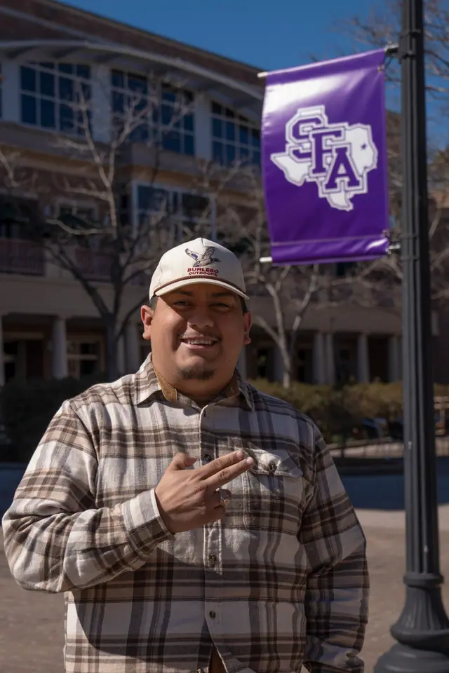 Student in a flannel shirt and cap on campus, a purple Stephen F. Austin banner on the lamppost behind him
