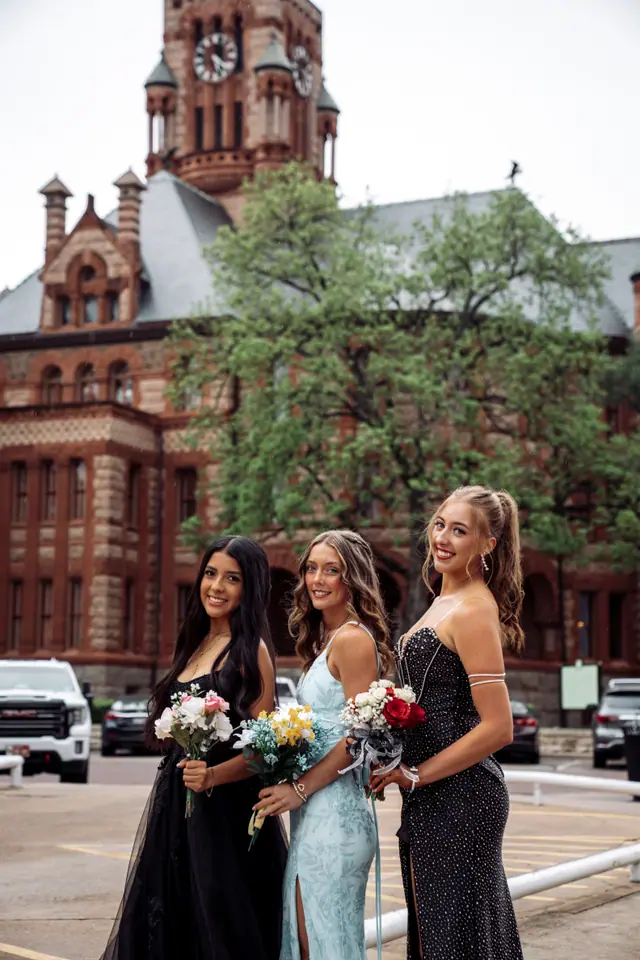 Three friends in prom gowns holding bouquets in front of a red brick historic courthouse