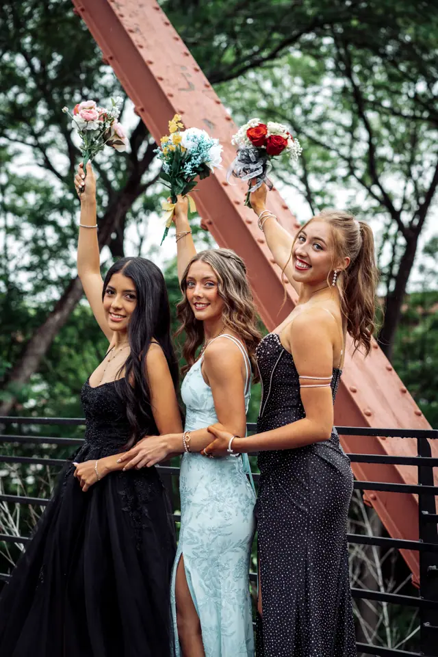 Three friends in prom gowns raising their bouquets beneath the red steel truss of a bridge