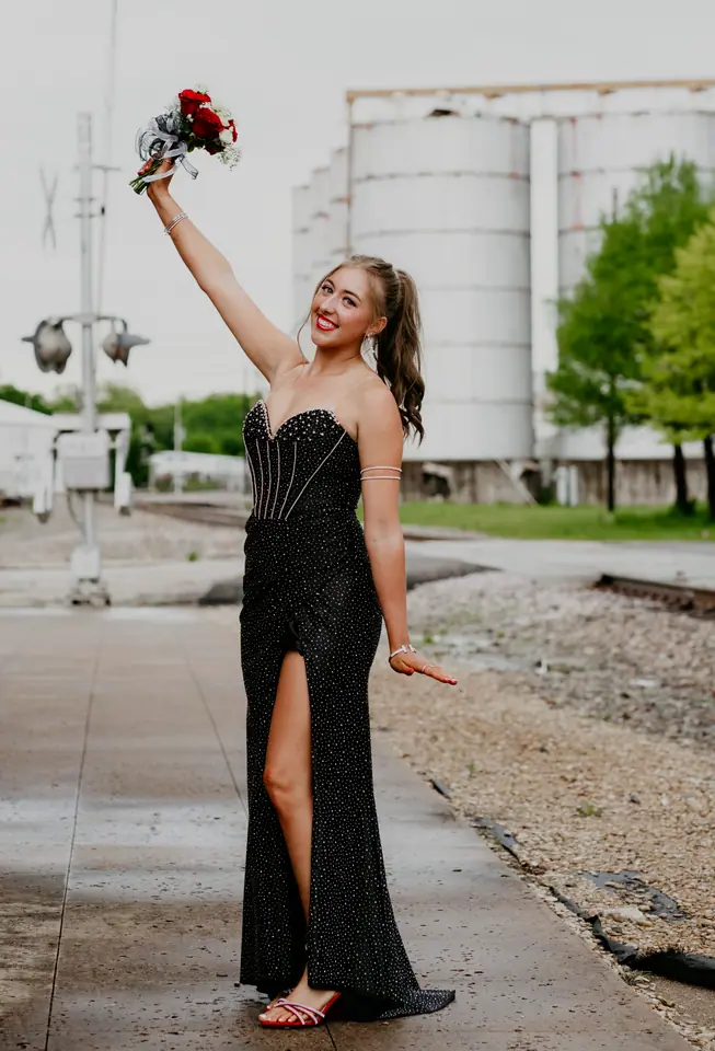 Young woman in a black beaded gown raising a rose bouquet overhead beside grain silos and a rail line