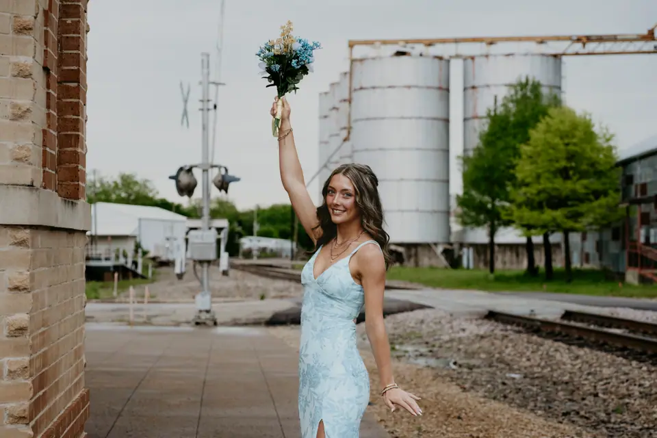 Young woman in a pale blue lace gown lifting a bouquet, brick building and grain silos behind her