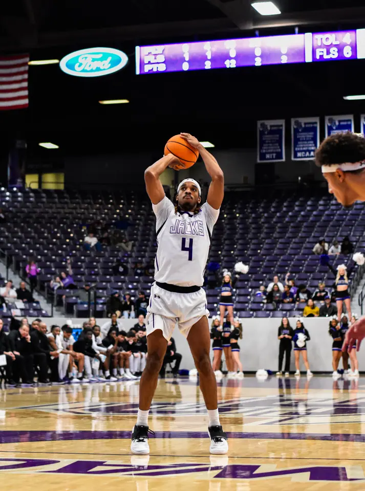 College basketball player rising for a jump shot in an arena, scoreboard lit above the stands